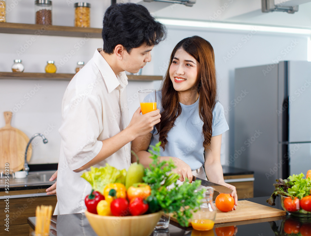 Asian young lovely couple handsome husband standing at kitchen counter full of fresh fruits and vegetables holding glass of orange juice ready to drink while beautiful wife cutting tomato with knife