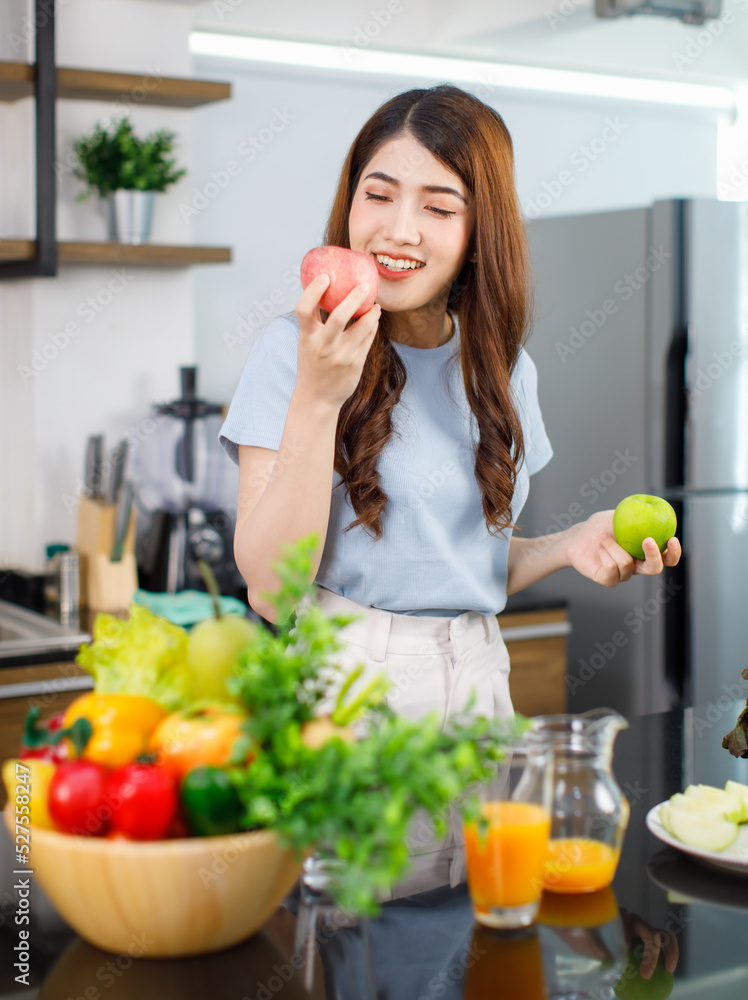 Asian young beautiful housewife in stripe apron standing smiling at kitchen counter full of organic fresh fruits and vegetables in bowls preparing salad peeling lettuce with hands on chopping board