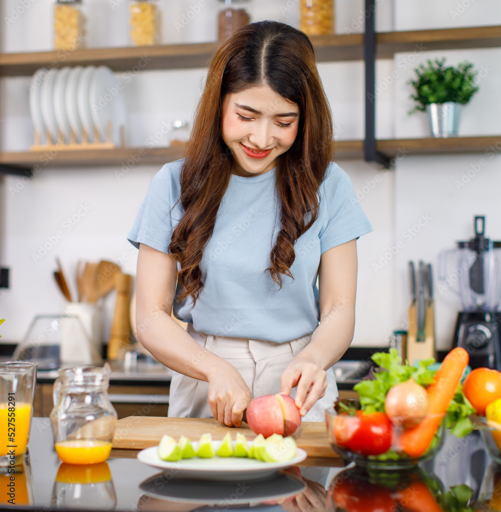 Asian young beautiful housewife standing at kitchen counter full of organic fresh fruits and vegetables bowl using knife preparing cutting red and green apple on chopping board ready to serve on dish