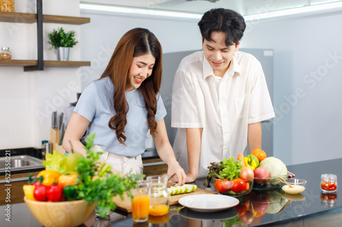 Asian young lovely couple beautiful wife at kitchen counter full of organic fresh fruits orange juice and vegetables using knife cutting green apple on chopping board while husband standing beside