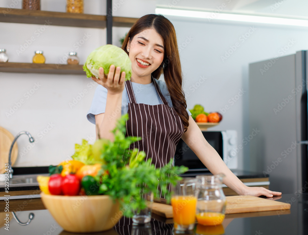 Asian young beautiful housewife in stripe apron standing smiling at kitchen counter full of organic fresh fruits and vegetables in bowls preparing salad peeling lettuce with hands on chopping board Asian young beautiful housewife in stripe apron standing smiling at kitchen counter full of organic fresh fruits and vegetables in bowls preparing salad peeling lettuce with hands on chopping board