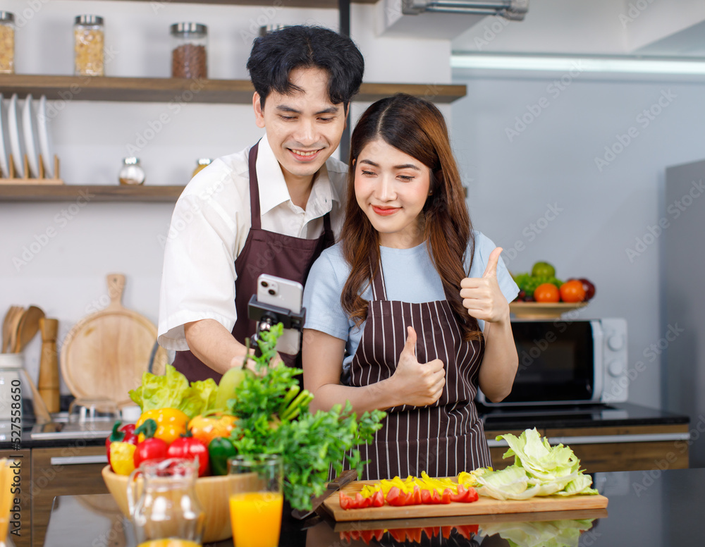 Asian millennial young lover couple handsome husband and pretty wife in apron standing smiling using selfie stick holding smartphone taking photo together in kitchen while preparing vegetables salad