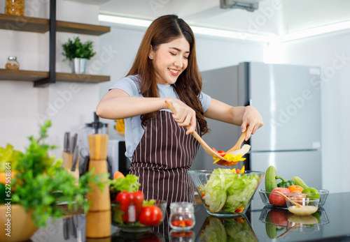 Asian young cheerful beautiful housewife in stripe apron standing smiling at kitchen counter using wood spoon and fork preparing mixing organic fresh mixed salad vegetables in glass bowl for dinner