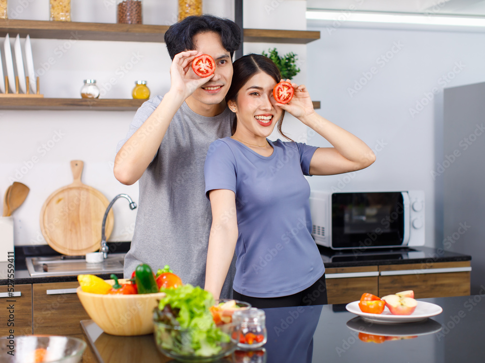 Asian young romantic couple handsome husband and beautiful wife standing smiling together behind kitchen counter full of fresh raw organic vegetables in glass bowl holding sliced red tomatoes on eyes Asian young romantic couple handsome husband and beautiful wife standing smiling together behind kitchen counter full of fresh raw organic vegetables in glass bowl holding sliced red tomatoes on eyes
