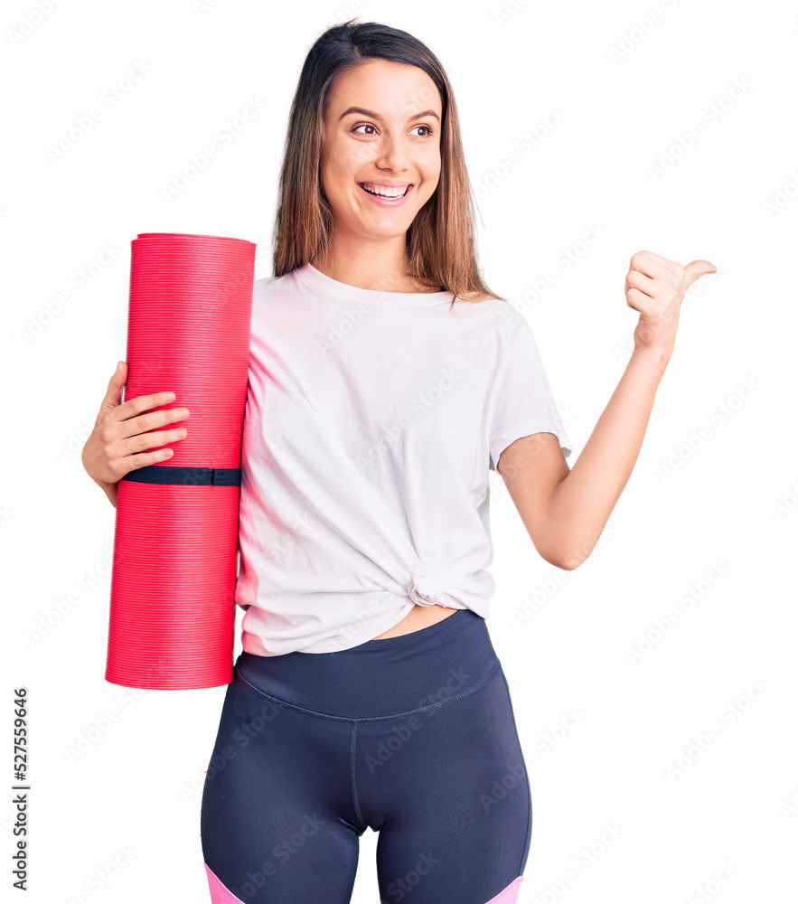 Young beautiful girl holding yoga mat pointing thumb up to the side smiling happy with open mouth Young beautiful girl holding yoga mat pointing thumb up to the side smiling happy with open mouth