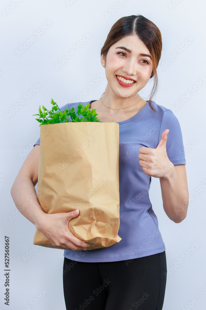 Asian young beautiful housewife in casual wear standing smiling showing and holding brown paper shopping bag of fresh raw organic vegetables from market on white background. Asian young beautiful housewife in casual wear standing smiling showing and holding brown paper shopping bag of fresh raw organic vegetables from market on white background.