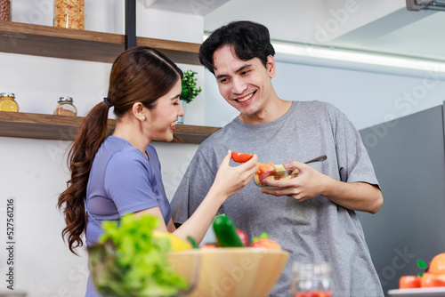 Asian young romantic couple beautiful wife standing smiling feed sliced mixed fruits from glass bowl to handsome husband behind kitchen counter full of fresh raw organic vegetables