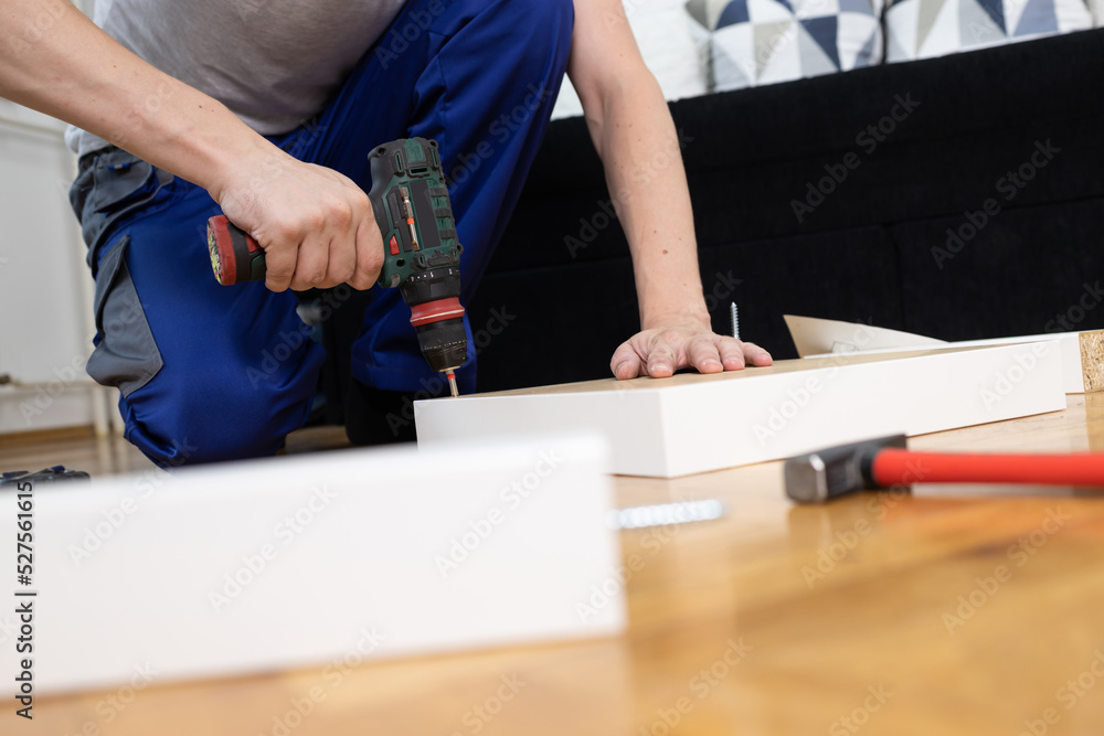 Male carpenter using electric drill assembling wooden table. Man In blue coverall uniform fixing desk with carpentry tools.
