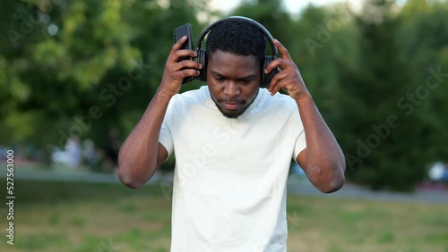 African American man putting on headphones walks in green park. Black guy types messages to mates on smartphone enjoying healthy trip photo