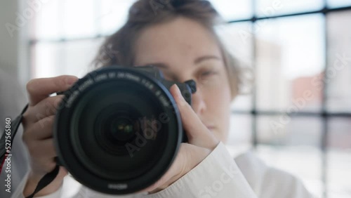 Closeup of young professional photographer woman ready to take picture with DSLR camera in photo studio with natural window daytime light on background. Process of blogging or hobby photography