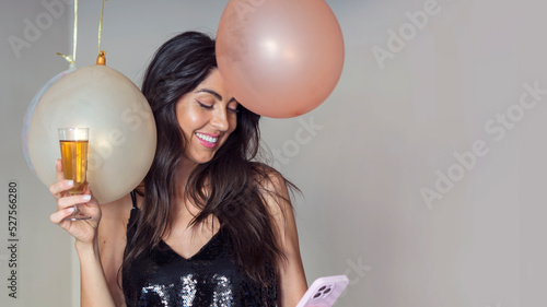 Beautiful young woman with shiny dress holding glass of wine and mobile phone on balloons background .Party time concept . Copy space 