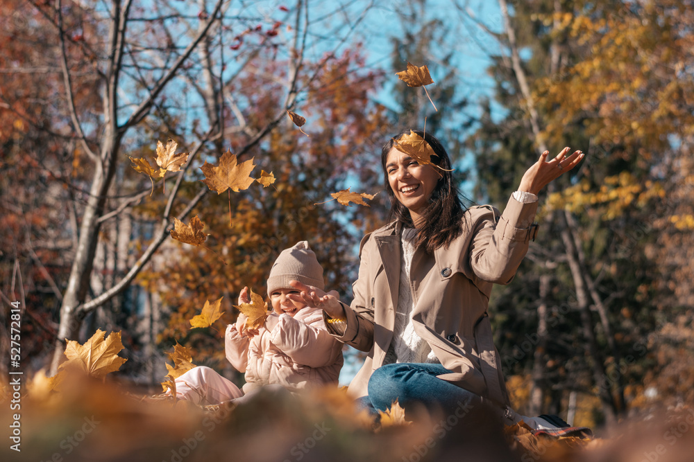 Happy dark-haired young mother and daughter having fun in the autumn park, throwing yellow leaves on a sunny day.Family and autumn concept.