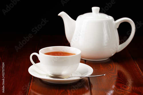 White ceramic cup of tea with a saucer and a spoon on a black background. In the background is a white porcelain teapot. Side view. Selective focus.