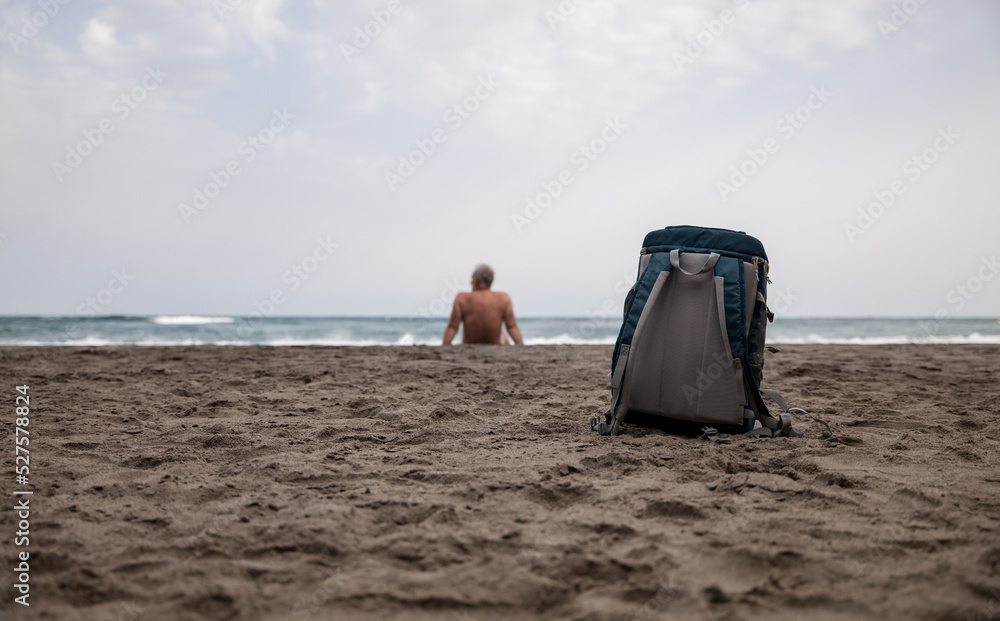 Backpack on beach with a shirtless man in background against sea and sky