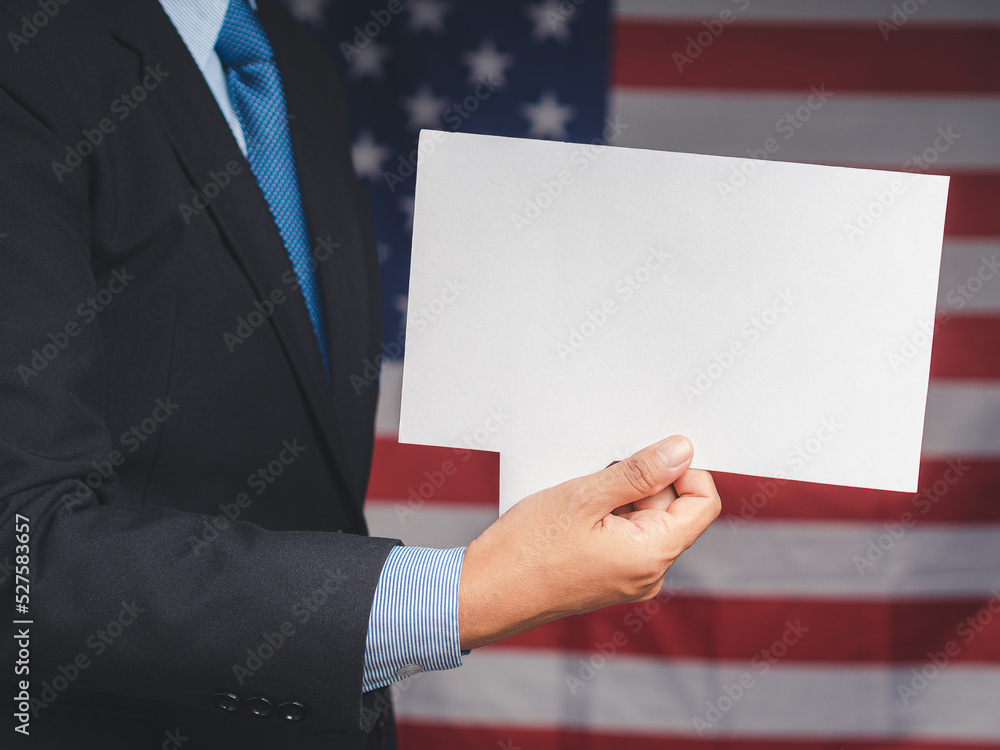 Close-up of a hand businessman holding a blank white speech bubble while standing on the American flag background Close-up of a hand businessman holding a blank white speech bubble while standing on the American flag background