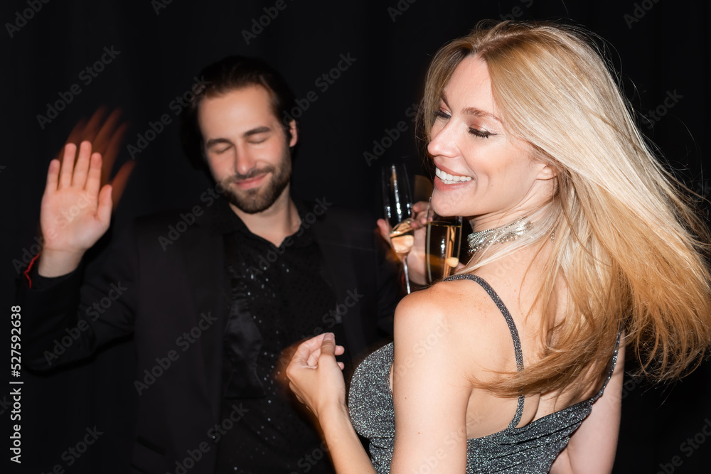 Motion blur of smiling woman with champagne dancing with boyfriend isolated on black.