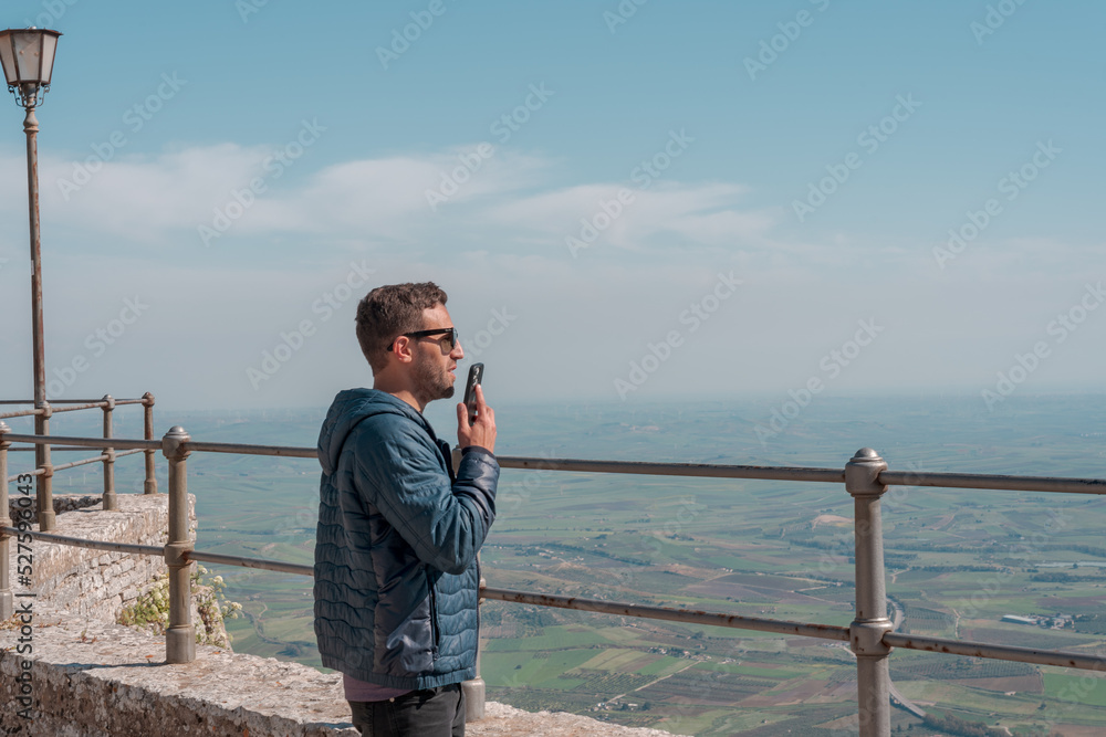 Smiling young man with wearing casual clothes over beautiful landscape background sending voice message on her smart phone. Communication and new technologies concept. Smiling young man with wearing casual clothes over beautiful landscape background sending voice message on her smart phone. Communication and new technologies concept.
