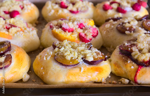 Closeup of freshly baked homemade sweet rolls with various seasonal fruits (strawberries, nectarines, plums, bananas) and golden crumble. The rolls are placed on a baking tray lined with paper.