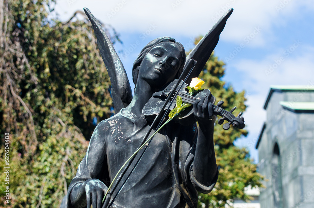 La statua bronzea di un angelo che suona il violino su cui hanno appoggiato una rosa gialla su una tomba dell cimitero maggiore di Milano