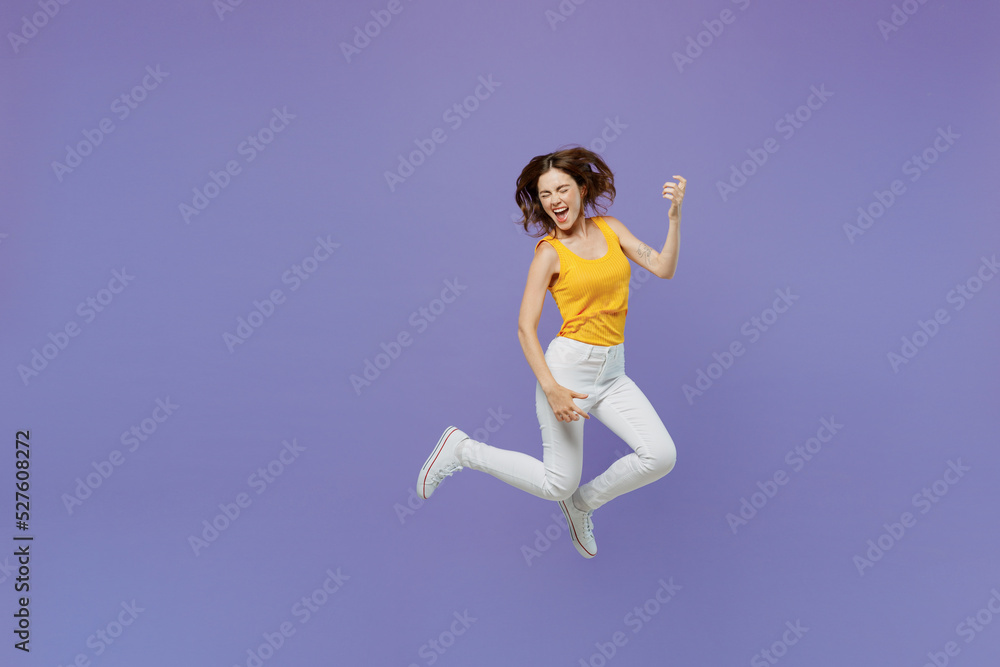 Full body expressive singer happy young woman she wear yellow tank shirt jump high play guitar hand gesture isolated on plain pastel light purple background studio portrait. People lifestyle concept.