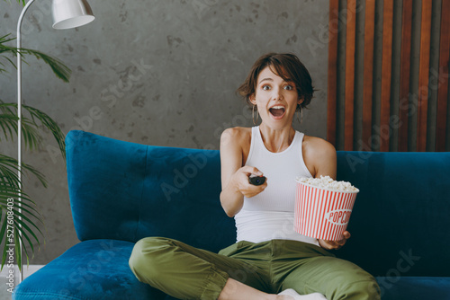 Young shocked woman in white tank shirt hold TV remote controller pop corn watch film switch channels sit on blue sofa stay at home hotel flat rest relax spend free spare time in living room indoors