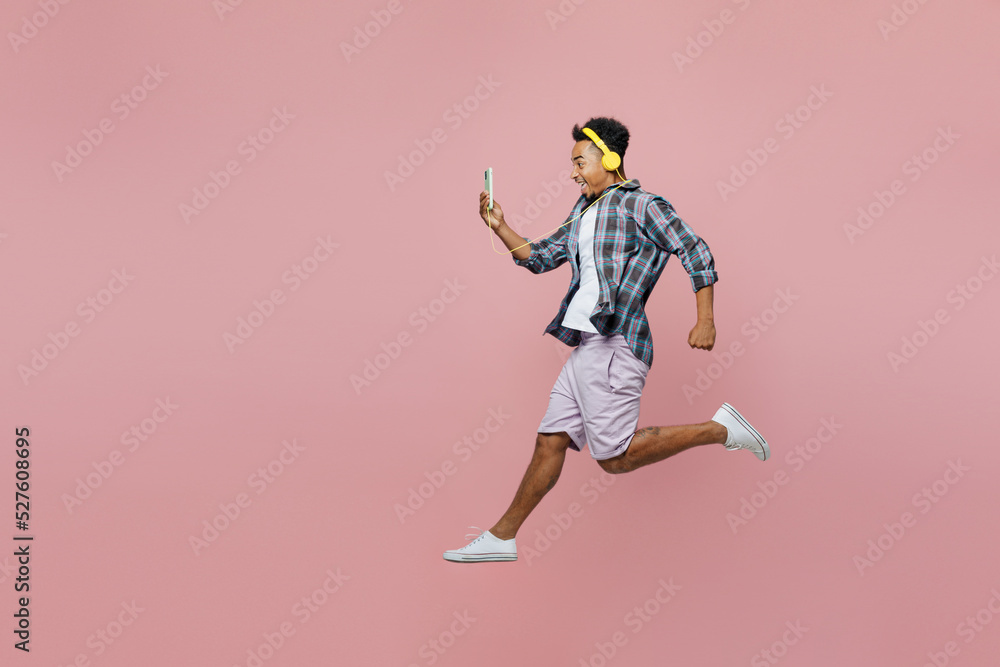 Full body side view young excited man of African American ethnicity 20s wear blue shirt use mobile cell phone listen music in headphones isolated on plain pastel light pink background studio portrait.