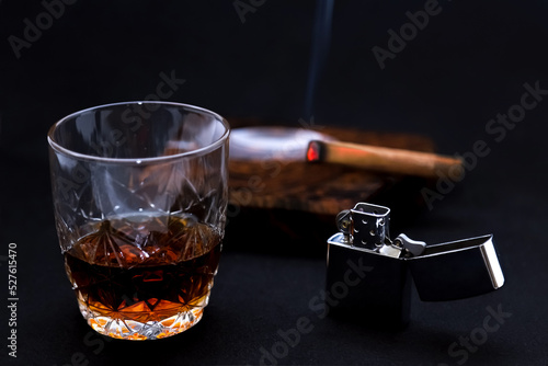 An antique lighter with a glass of brandy on the background of a blurred smoking cigar. Shallow depth of field