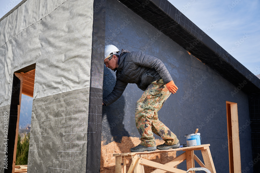 Male painter using paint roller, doing exterior paint work in a black color. Man worker building wooden frame house. Carpentry and construction concept. Male painter using paint roller, doing exterior paint work in a black color. Man worker building wooden frame house. Carpentry and construction concept.
