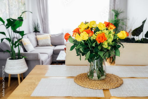 Kitchen counter table with focus on vase with yellow and orange roses on it with blurred background of modern cozy living room with couch and green plants. Open space home interior design. Copy space.