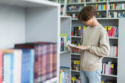 teenage student reading book near bookshelves in library on blurred foreground.