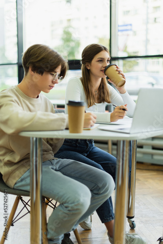 teenage girl drinking coffee near laptop and friend writing in library reading room.