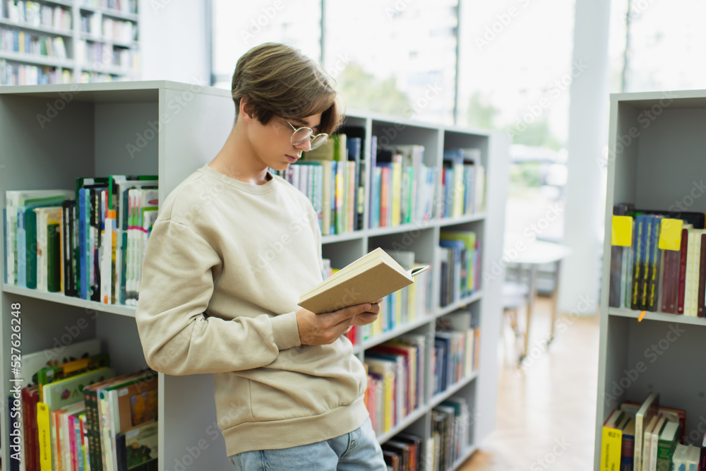 student in eyeglasses reading book while standing near shelves in library. student in eyeglasses reading book while standing near shelves in library.