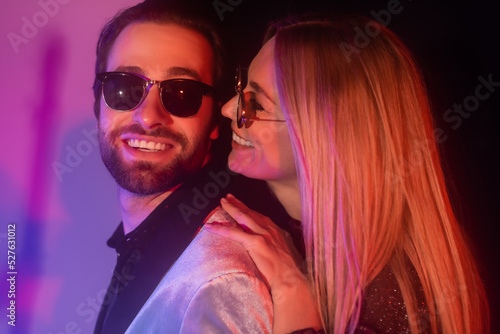 Cheerful blonde woman in sunglasses hugging boyfriend in jacket on purple background.
