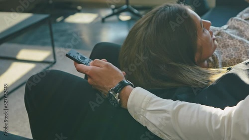 Man in white shirt, vest, sits leather sofa after hard day work. Switches TV channels streaming service from remote, smiles. Woman pajamas lies her husband lap, smiles, flirts. Rays setting sun wall