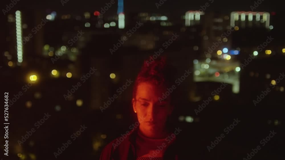 Portrait of young woman posing for camera in flickering red light on dark street with night city in background