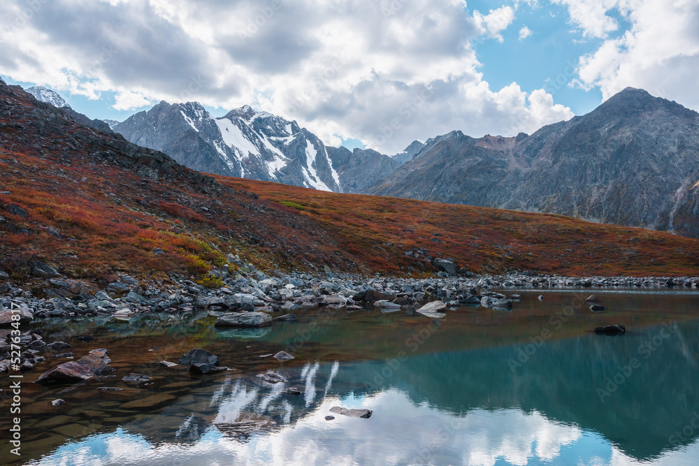 Lovely autumn landscape with pure turquoise mountain lake and red hill against high snowy mountain range under clouds in blue sky. Beautiful alpine lake and large snow mountains. Fading autumn colors.