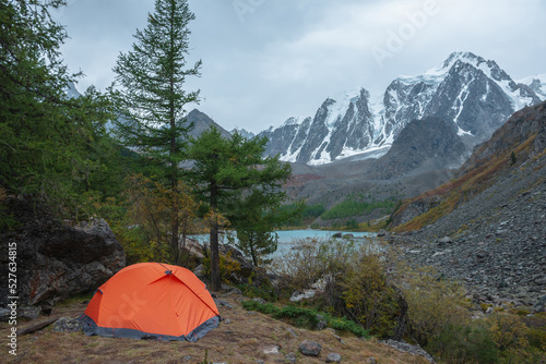 Dramatic landscape with alone orange tent on forest hill among rocks and autumn flora with view to large snowy mountain range under cloudy sky. Lonely tent and fading autumn colors in high mountains.