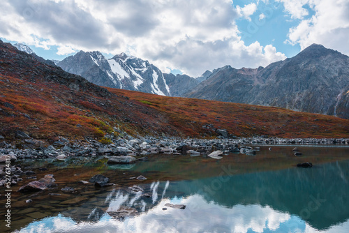 Lovely autumn landscape with pure turquoise mountain lake and red hill against high snowy mountain range under clouds in blue sky. Beautiful alpine lake and large snow mountains. Fading autumn colors.