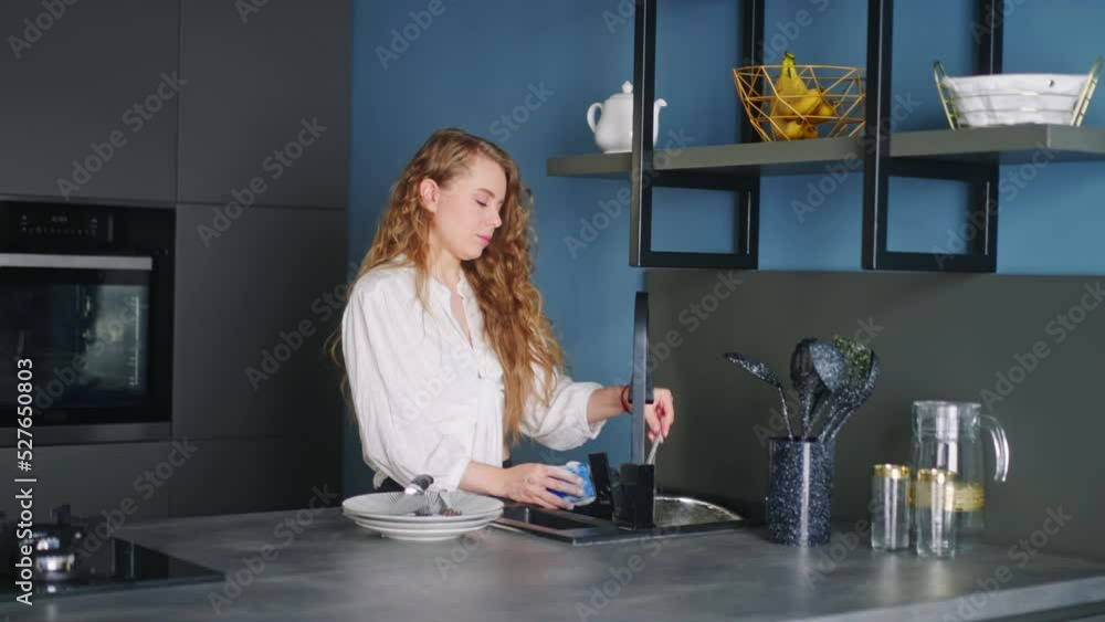 Young caucasian woman washes spoon, fork and knife under running water in sink in modern kitchen. Housewife is rinsing cutlery on kitchen island. Female performing the washing-up in high-tech kitchen Young caucasian woman washes spoon, fork and knife under running water in sink in modern kitchen. Housewife is rinsing cutlery on kitchen island. Female performing the washing-up in high-tech kitchen