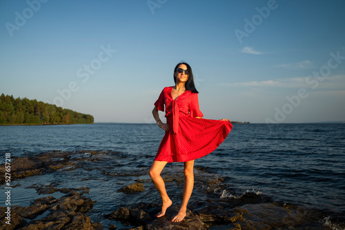 Slender brunette in sunglasses and a red dress posing on the shore