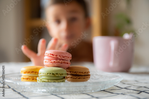 Child trying to catch a stack of delicious macaroons, of three colors, on a table with a checkered tablecloth, and a cup of coffee in the background