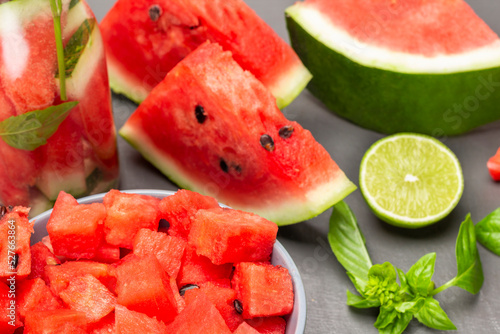 Sliced watermelon pulp in bowl. Slices of watermelon, half a lime and sprig of basil on table.