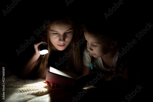 A teenage girl with her sister read in the dark with a flashlight.