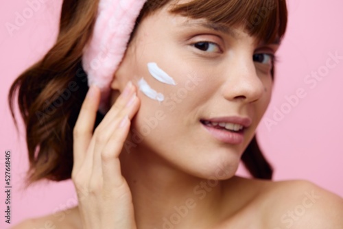 a woman with dark hair stands on a pink background in a white towel with a pink bandage on her head with a cream applied to her cheek, takes care of her skin. Horizontal studio photography.