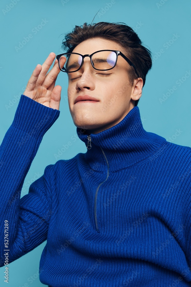 attractive man stands on a light blue background in a blue zip-up sweater and holding black-rimmed glasses on his face with his hand, closed his eyes while biting his lower lip. Vertical Photography attractive man stands on a light blue background in a blue zip-up sweater and holding black-rimmed glasses on his face with his hand, closed his eyes while biting his lower lip. Vertical Photography