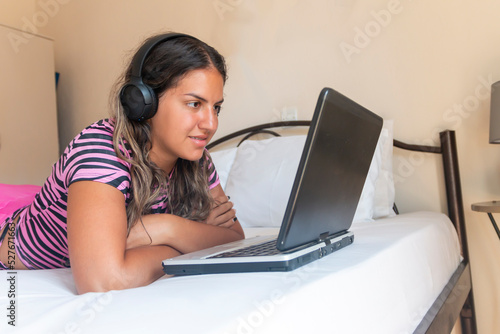 Teenager girl resting in bed, listening to music using computer, enjoying communicating in social network, web surfing information, shopping online or e-dating. © Jelena