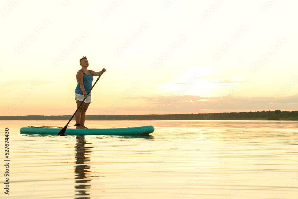 A woman in mohawk shorts stands on a SUP board at sunset in a lake against a pink-blue sky and water.