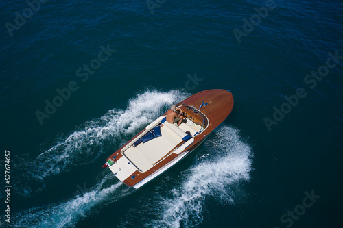 Italian wooden speedboat moving up aerial view. Wooden expensive boat fast movement on dark water top view. Big expensive wooden open boat with a man moving fast on dark blue water top view. © Berg