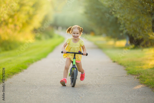 Happy beautiful little girl fast running and riding on first bike without pedals on sidewalk at city park in warm summer day. Cute 3 years old toddler. Front view. Learning to keep balance. © fotoduets