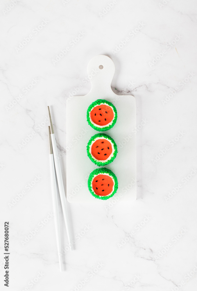 Mousse cream cakes with jelly watermelon in the form of round sweet Japanese sushi, sprinkled with green coconut chips, on a serving board. Light background. Top view Mousse cream cakes with jelly watermelon in the form of round sweet Japanese sushi, sprinkled with green coconut chips, on a serving board. Light background. Top view